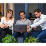 Three people sit comfortable on a couch and look at a laptop screen.