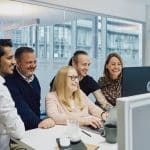 A group of people from Billwerk+ stand in front of a standing desk and look at the computer screen. Everyone is very happy.