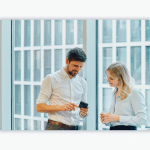 Two people stand at a large office window and look at a smartphone.