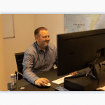 A man sits at his computer and smiles. He sits in front of a map of the world.