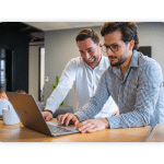 Two people are at a standing desk and look at a laptop.