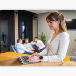 A woman works at a laptop at a standing desk. Three colleagues sit in the background and have a conversation